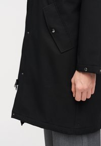 Πλευρική όψη ενός ατόμου που φοράει μαύρο παλτό με φερμουάρ και καπάκι τσέπης με κουμπιά, και γκρίζο παντελόνι, με το χέρι χαλαρό στο πλάι.