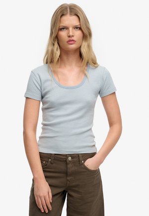 Blondynka z rozpuszczonymi włosami, ubrana w jasnoniebieski obcisły top w paski z krótkim rękawem oraz ciemnobrązowe spodnie, z prawą ręką w kieszeni.