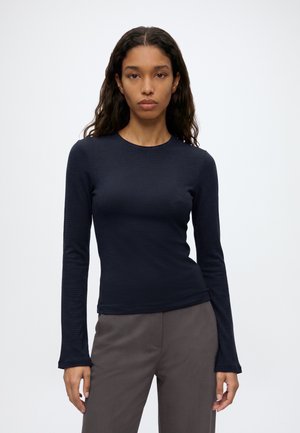 Femme aux longs cheveux foncés, portant un haut ajusté à manches longues bleu marine et un pantalon gris, debout devant un fond clair uni.