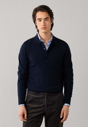 Jonge man met donker haar die een marineblauw poloshirt met lange mouwen draagt over een lichtblauw overhemd met kraag en een donkere broek tegen een effen achtergrond.