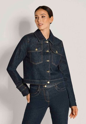 Frau trägt dunkelblaue Jeansjacke und passende Jeans, posiert mit einer Hand in der Tasche vor schlichtem hellem Hintergrund.