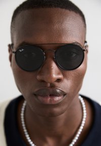 Round black sunglasses with a thin metal frame, featuring dark lenses. A slight reflection is visible. The wearer has smooth skin and neutral lips.