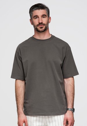 Mann mit kurzem dunklem Haar und Bart trägt ein lockeres dunkelgraues T-Shirt und weiße gestreifte Shorts, stehend vor einem schlichten hellen Hintergrund.