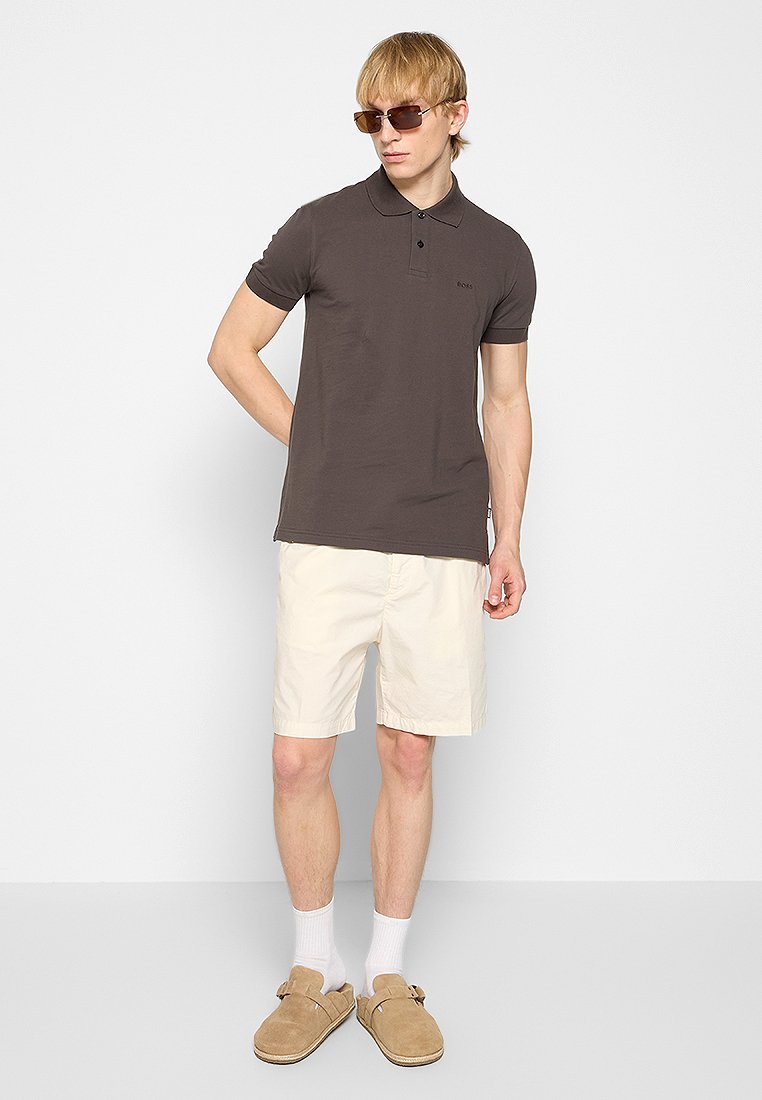 Jeune homme portant un polo marron foncé, un short crème, des chaussettes blanches, des chaussures à enfiler beiges et des lunettes de soleil marron, le tout sur un fond clair et uni.