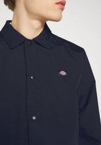 Jeune personne portant une veste noire boutonnée avec un petit logo de la marque Dickies sur la poitrine gauche, vue de profil.
