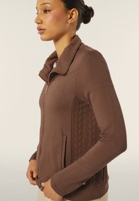 Giacca marrone con zip, caratterizzata da un collo testurizzato, pannello laterale con motivo trapuntato e tasche frontali. Tessuto liscio per una vestibilità comoda.