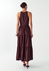 Nainen, jolla on bun-mallinen kampaus, pukeutuneena hihattomaan tummanruskeaan kerrokselliseen maksimekkoon, mustiin korkokenkiin, kultaisiin renkaisiin ja paksuihin rannekoruihin.