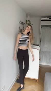Žena s dlouhými blond vlasy, oblečená v černobílém pruhovaném crop topu a černých kalhotách, stojící a opírající se o bílou stěnu uvnitř.