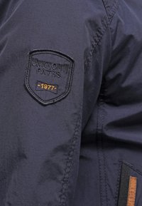 Marineblauwe jas met een gestructureerd oppervlak, voorzien van een geborduurd logo met de tekst "UNKNOWN PATHS" en "1977" in bruin.