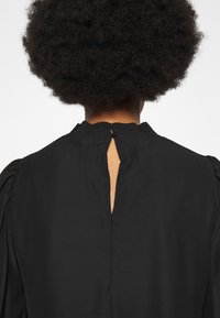 Blouse noire avec un col froncé et une fente à l'arrière. Texture lisse avec des manches bouffantes et sans motifs visibles.