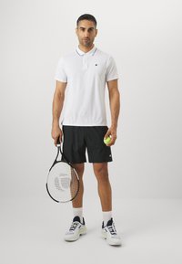 Camisola polo branca com detalhes em preto no colarinho, calções pretos, segurando uma raquete de ténis e uma bola de ténis, usando sapatilhas de desporto e meias brancas.