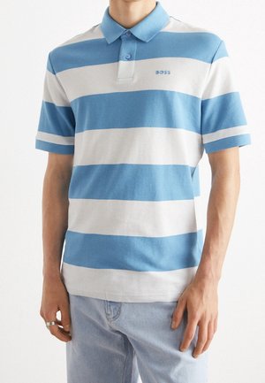 Man draagt een kortarmig poloshirt met brede horizontale blauwe en witte strepen en lichtblauwe spijkerbroek, staand met handen ontspannen langs het lichaam.