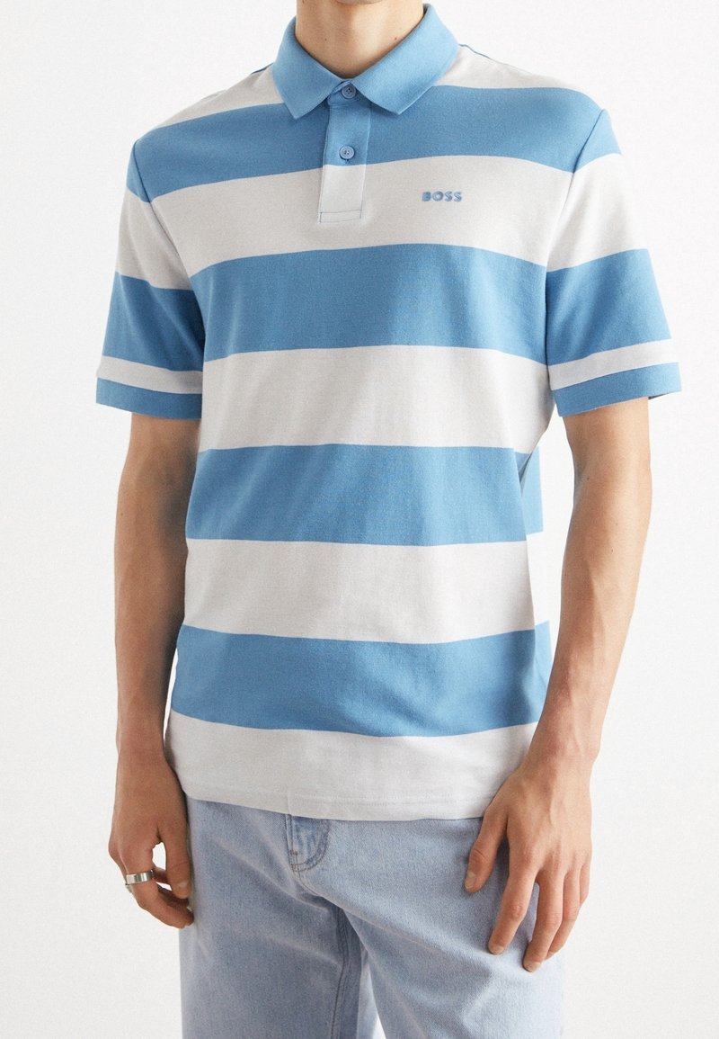 Homme portant un polo à manches courtes avec de larges rayures horizontales bleues et blanches et un jean bleu clair, debout, les mains détendues le long du corps.