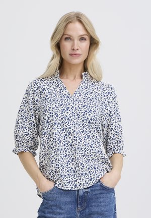 Femme aux cheveux blonds longs, portant un chemisier bleu et blanc à motifs floraux et un jean bleu, debout les mains dans les poches sur un fond blanc.