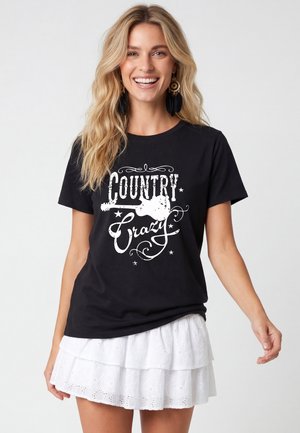 Uśmiechnięta kobieta o blond włosach, ubrana w czarną koszulkę "Country Crazy" i białą warstwową spódnicę, stojąca na jednolitym jasnym tle.