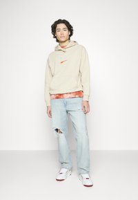 Kremhet genser med PUMA-logo, lagd over en oransje tie-dye skjorte. Brukt sammen med lyseblå slitte jeans og hvite sneakers med røde detaljer.