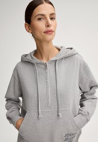 Szara bluza z kapturem na zamek, wykonana z miękkiego materiału, z przednią kieszenią typu kangur i regulowanymi sznurkami na kapturze. Nadruk z logo na kieszeni.