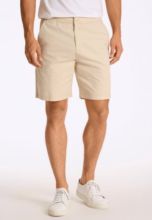 Mand iført beige shorts, hvid T-shirt og hvide sneakers står foran en ensfarvet lys baggrund.
