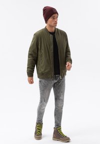 Giacca bomber verde oliva con polsini a costine, t-shirt nera, jeans grigi strappati e stivali marroni con lacci verde fluorescente.