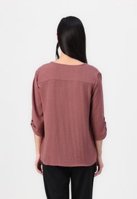 Blusa di un colore ruggine, con texture, maniche a tre quarti e polsini arrotolati. Collo rotondo, vestibilità morbida e orlo leggermente curvato.