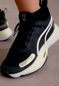 Schwarzer Sportschuh mit Obermaterial aus Mesh, weißen Akzentstreifen, strukturiertem Sohlenprofil und reflektierenden Details. Entwickelt für Komfort und Unterstützung.