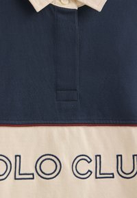 Kurzärmeliges Poloshirt in Marineblau und Creme, mit Kragen, Knöpfen und der blauen Stickerei "POLO CLUB" auf dem cremefarbenen Bereich.
