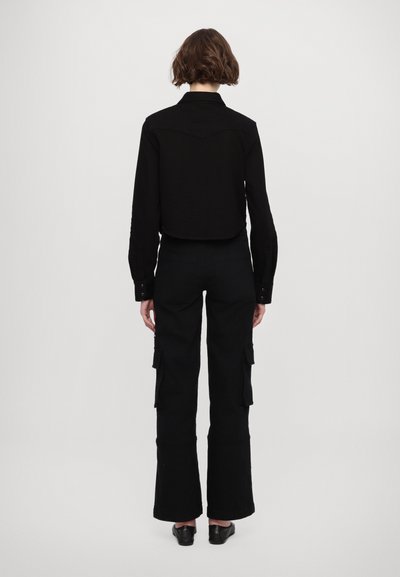 Cropped shirt nera con colletto abbinata a pantaloni cargo neri a vita alta, dotati di tasche laterali e gamba leggermente svasata.