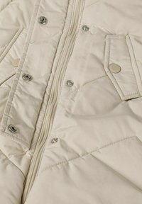 Beige gewatteerde pufferjack met zilveren drukknoopjes en een rits, met een gladde textuur en subtiele glans. Voorzien van twee voorzakken.