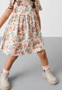 Robe à imprimé floral en crème avec des fleurs roses et bleues, manches courtes et taille froncée. Bottines beige à la cheville avec fermeture éclair sur le côté et semelles texturées.
