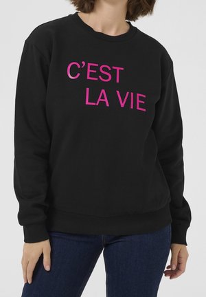 Personne portant un sweat-shirt noir avec un texte rose indiquant ""C'EST LA VIE"" et un jean bleu sur un fond uni.