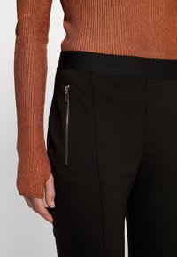 Pantaloni neri elasticizzati con tasca laterale con zip e una cintura elastica liscia. La parte superiore ha una vestibilità aderente con texture a righe arancioni.
