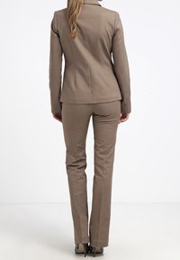 Na míru šitý béžový oblek se přiléhavým blejzrem a kalhotami rovného střihu. Hladká textura, jednokapsový vent, a elegantní design. Na viditelných podpatcích.