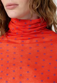 Sieviete, kas valkā spilgti oranžu turtleneck džemperi ar purpura punktiņiem, atklājot savu kaklu, lūpas un viļņainus gaiši brūnus matus.