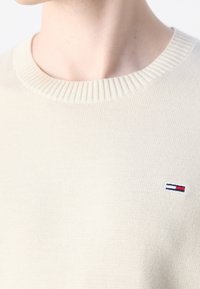 Gros plan d'une personne portant un pull en maille crème avec un petit logo Tommy Hilfiger sur la poitrine.