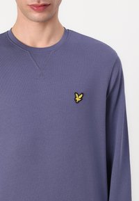 Sweatshirt violet à manches longues en tissu doux, doté d'un col rond et d'un petit logo d'aigle brodé jaune sur la poitrine gauche.