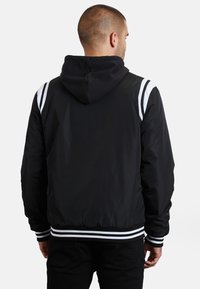 Schwarze Bomberjacke mit Kapuze, ausgestattet mit weißen Streifen auf den Schultern und gestreiften Bündchen. Glattes Material mit entspannter Passform.