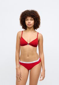 Punane puuvillane bralette õhukeste reguleeritavate rihmadega, millel on väike keskne hardware detail. Sobivad punased bikiinipüksid valge logo vööga.