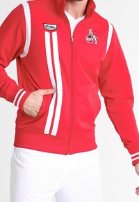 Veste zippée rouge avec des bandes blanches sur les épaules et les manches, avec un patch de marque sur la poitrine gauche et des poches latérales.