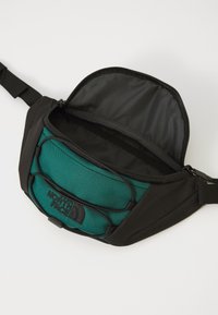 Fekete és türkiz fanny pack, cipzáras fő rekesszel, hálós részletekkel, állítható pánttal és elülső logóval.
