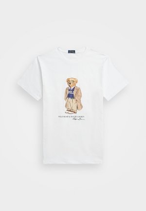 T-shirt en coton blanc avec un ours en dessin animé portant des lunettes de soleil, un blazer et un pull violet "Polo". Comprend une signature en dessous du design.