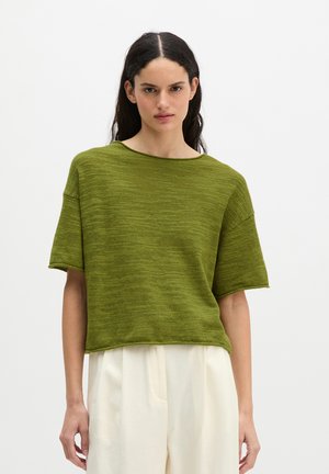 Femme aux longs cheveux foncés, portant un haut en maille ample à manches courtes vert olive et un pantalon blanc taille haute à jambes larges, debout devant un fond uni.