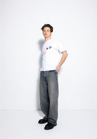 Biała koszulka z krótkim rękawem z graficznymi nadrukami, w połączeniu z wyblakłymi niebieskimi dżinsami w szerokim kroju i czarnymi klapkami na gumowej podeszwie.