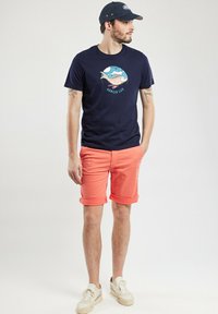 T-shirt en coton marine avec un graphique de poisson et un logo texte, assorti à un short corail à revers. Le modèle porte des baskets beiges et une casquette marine.