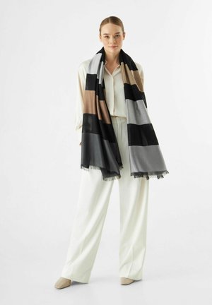 Écharpe rayée en tissu noir, gris, marron et beige avec des bords frangés. Portée drapée sur une tenue de couleur claire. Texture douce.