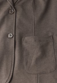 Vêtement marron à boutons, avec une texture lisse, présentant une poche poitrine et des détails de coutures visibles. Deux boutons sont affichés en évidence.