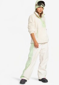 Fleece-pullover i creme med grønne detaljer, kombineret med hvide bukser med grønne sidelommer. Modellen bærer sorte snebriller og støvler.