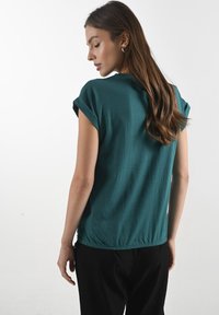 Žena s dlouhými, vlnitými vlasy má na sobě tyrkysovou košili a černé kalhoty, viděná zezadu na jednobarevném bílém pozadí.