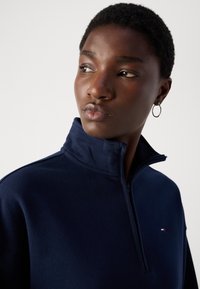 Navy blue zip-up sweatshirt featuring a high neck, smooth texture, and a small logo with red, white, and blue accents.
