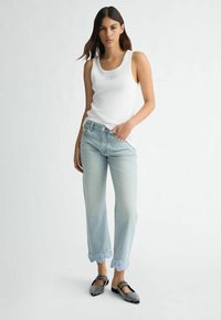 Débardeur blanc à côtes avec un petit logo, jeans bleus clairs avec un ourlet en dentelle brodée, et des chaussures plates noires à clous.