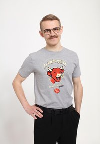Šedé bavlněné tričko s barevným potiskem červené krávy s náušnicemi a textem, zkombinované s černými kalhotami a černým opaskem.